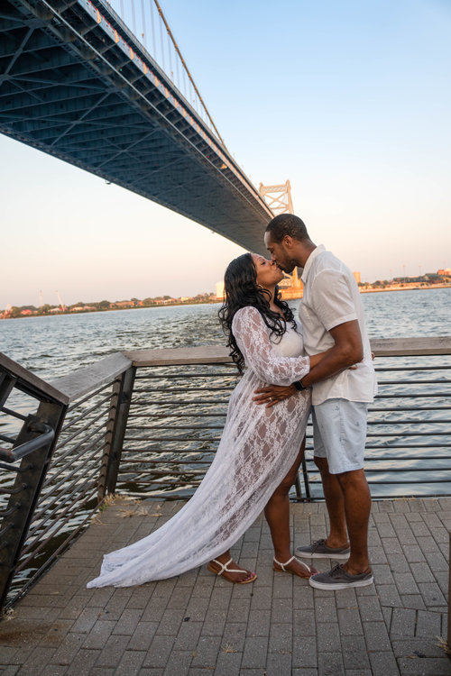 Couple kissing by a river under a blue bridge, with a city skyline in the background.