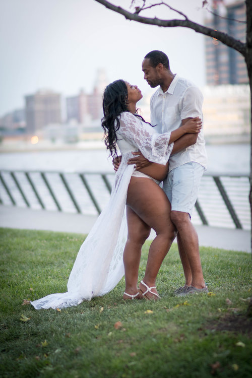 A couple embracing and smiling outdoors near a waterfront. The woman is wearing a sheer white robe and the man is in a casual white shirt and shorts. They are standing on a grassy area with trees and buildings in the background.