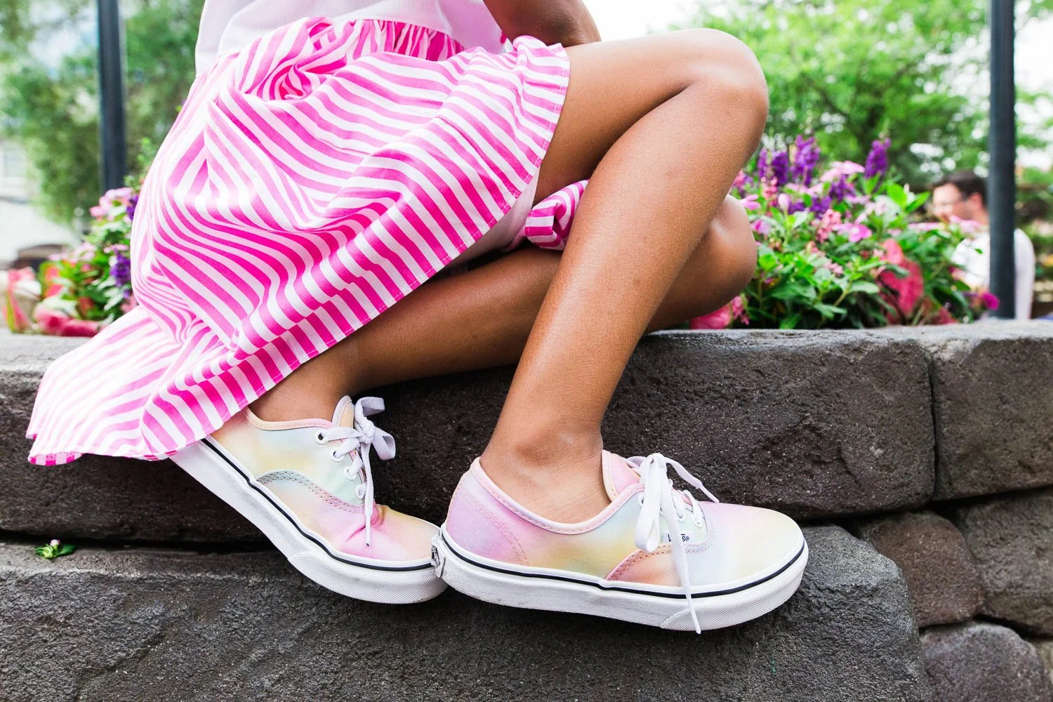 Little girl wearing pink striped dress and pink tie dye shoes sits on wall in Magic Kingdom.