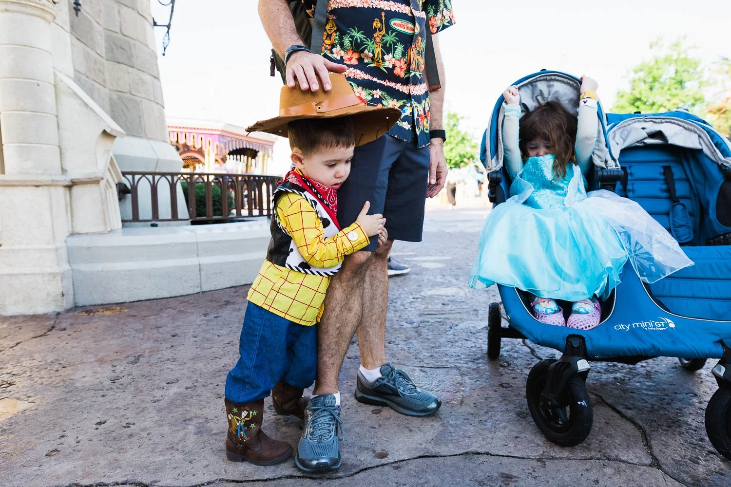 Little boy dressed like Woody holds onto his dad's leg in the Magic Kingdom.