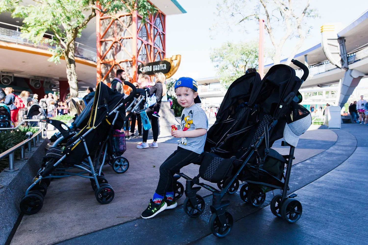 Boy sits on the edge of a stroller in Tomorrowland at the Magic Kingdom.