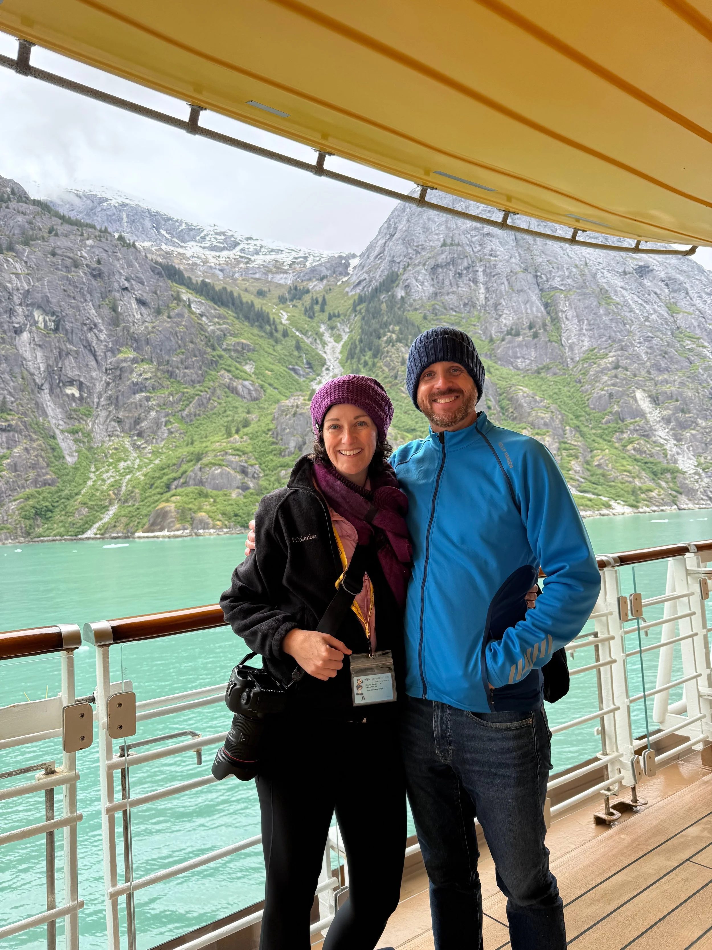Couple poses on deck four of the Disney Wonder with Alaska mountains in the background.