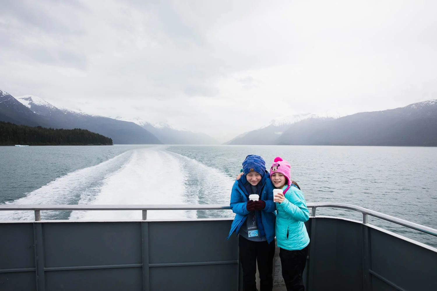 Siblings look tiny posing on the back of a boat in Alaskan wilderness.