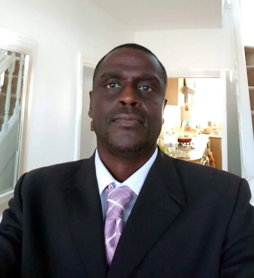 A man in a black suit and purple striped tie taking a selfie in a home interior, with a staircase and kitchen visible in the background.