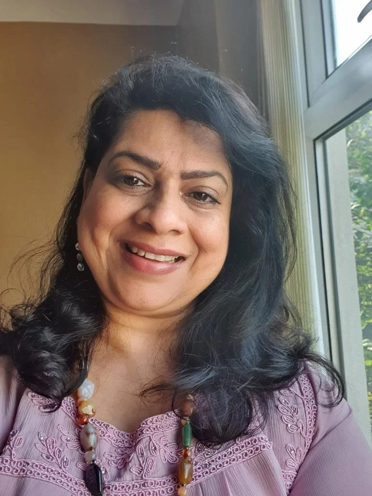 A woman with black wavy hair, wearing a pink embroidered top and a beaded necklace, smiling while taking a selfie near a window.
