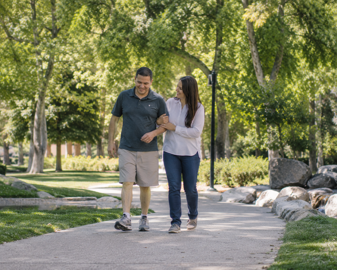 A couple walking together on a park path, smiling and engaging in conversation, surrounded by green trees and rocks.