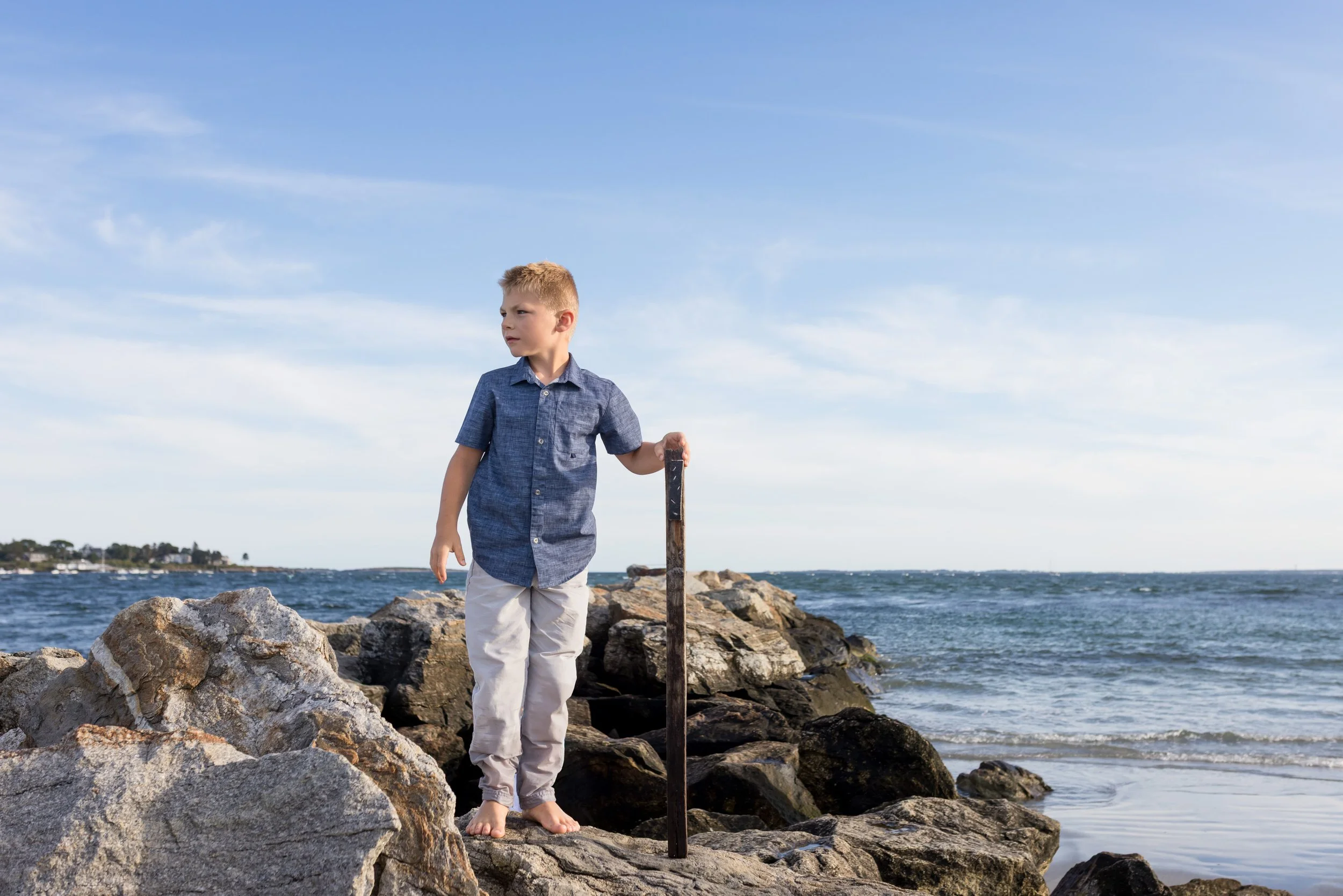 a boy standing on a rock wall, holding a stick, with the ocean behind him. He is looking off to the left of the camera.