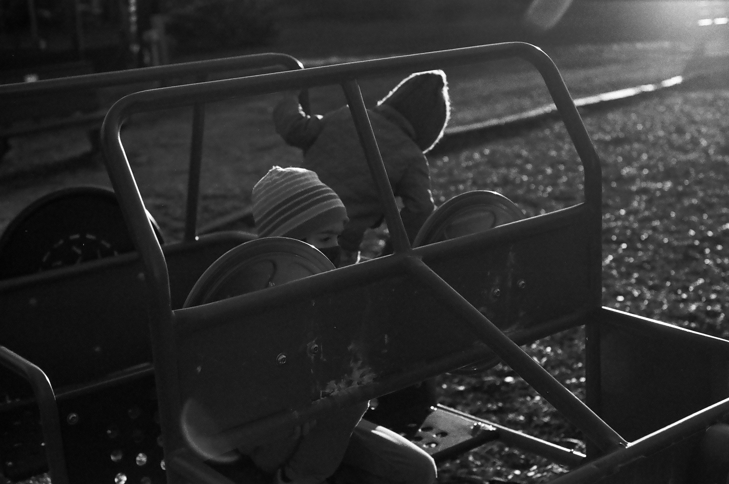 two children playing on playground equipment with the sun shining down on them. they are both wearing masks and hats.