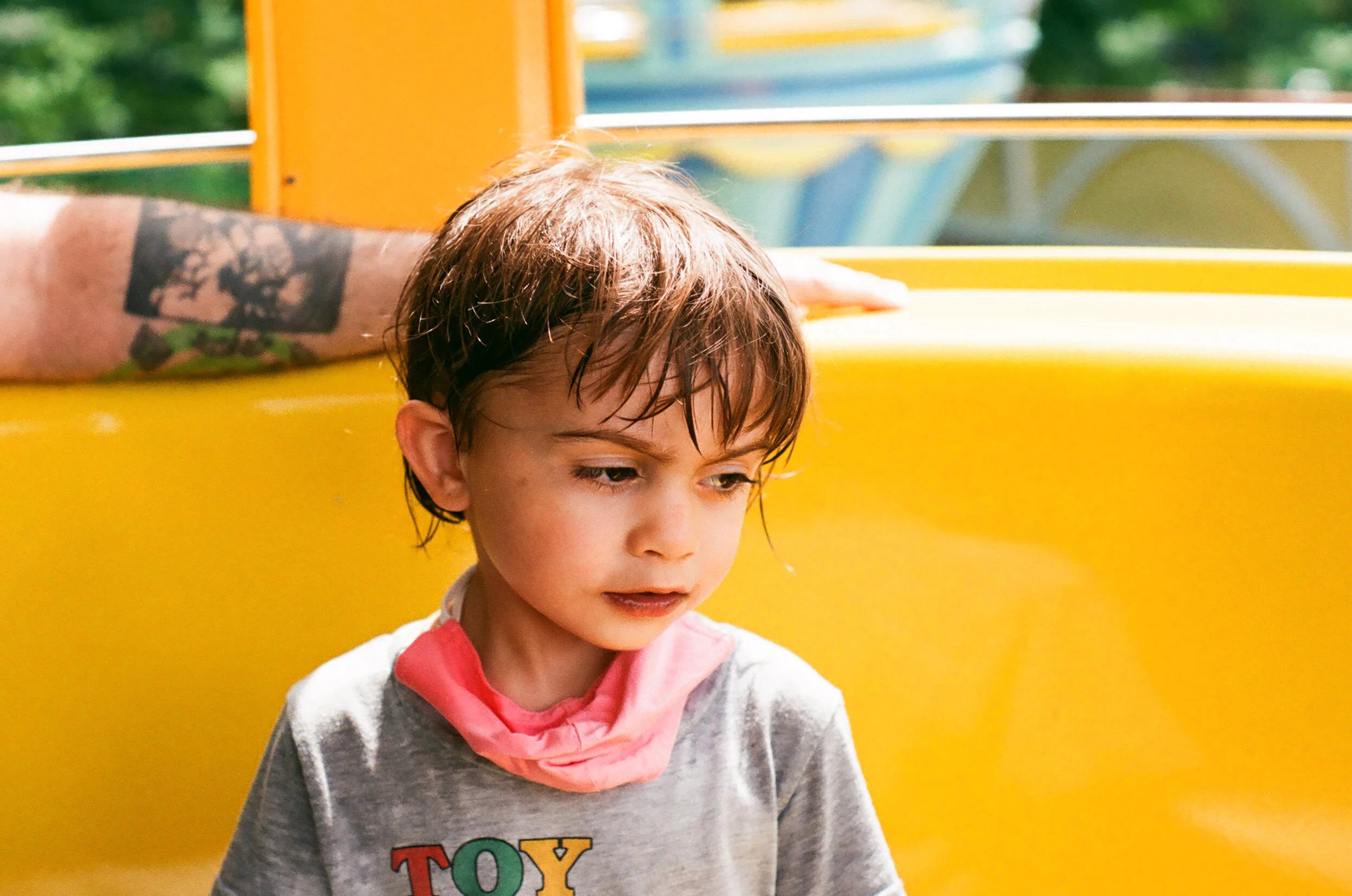 a small child with wet hair looks to the right of the camera, their brow is furrowed and they are looking down. the background is bright yellow. You can see part of someone's arm wrapped behind them.