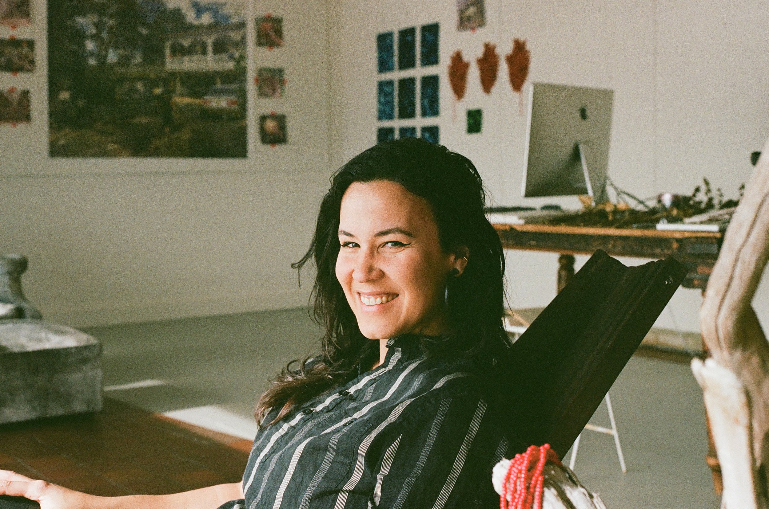  an image of artist Shey 'Rí Acu' Rivera Ríos (they/them) at Surf Point in York Maine. They are sitting in a chair, there is art hangin on gallery walls in the background. There is a piece of carved driftwood in the foreground right corner of the pho