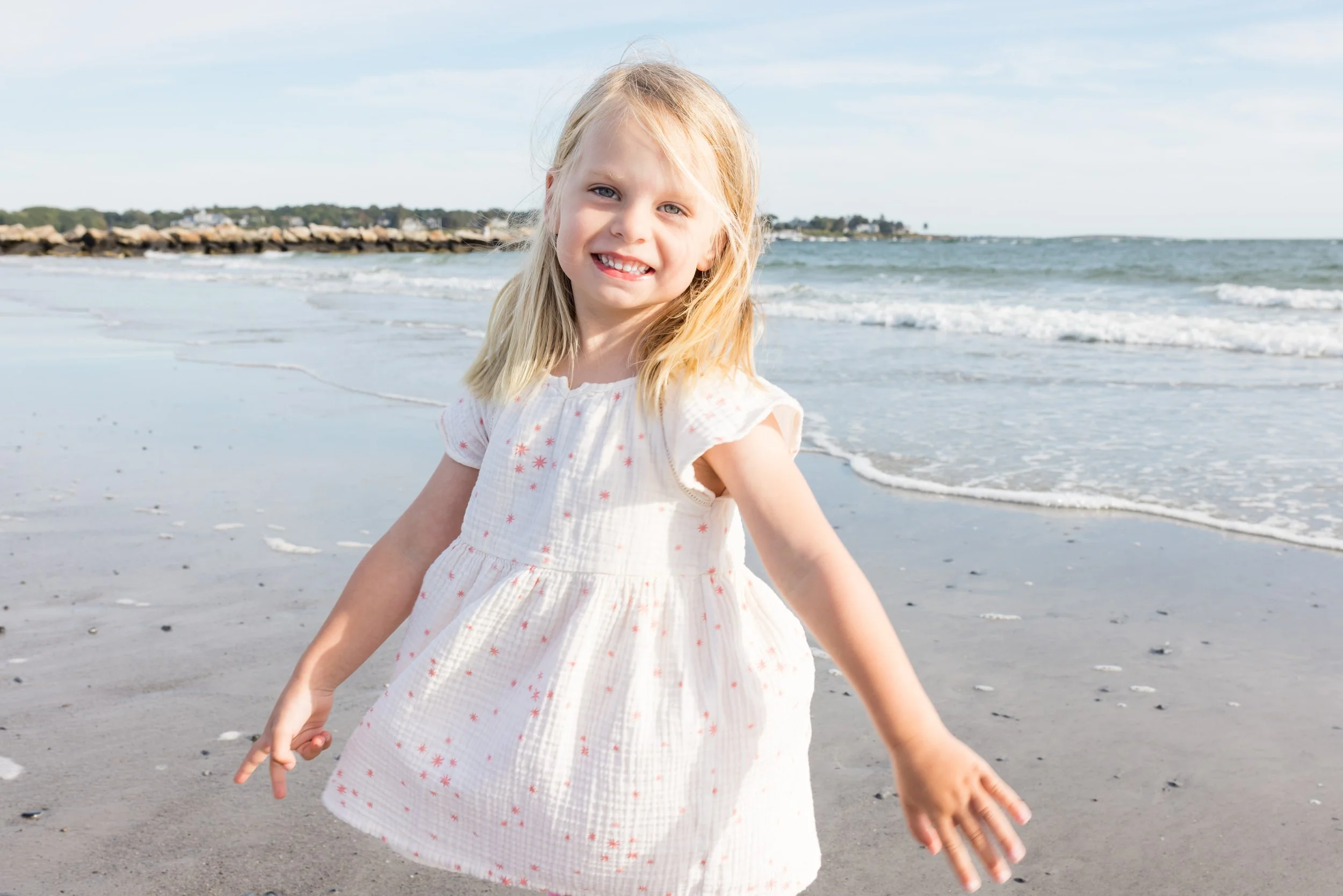 a girl with blond hair and blue eyes is looking at the camera, behind her is the beach and ocean. She is wearing a white top with little pink flowers on it.