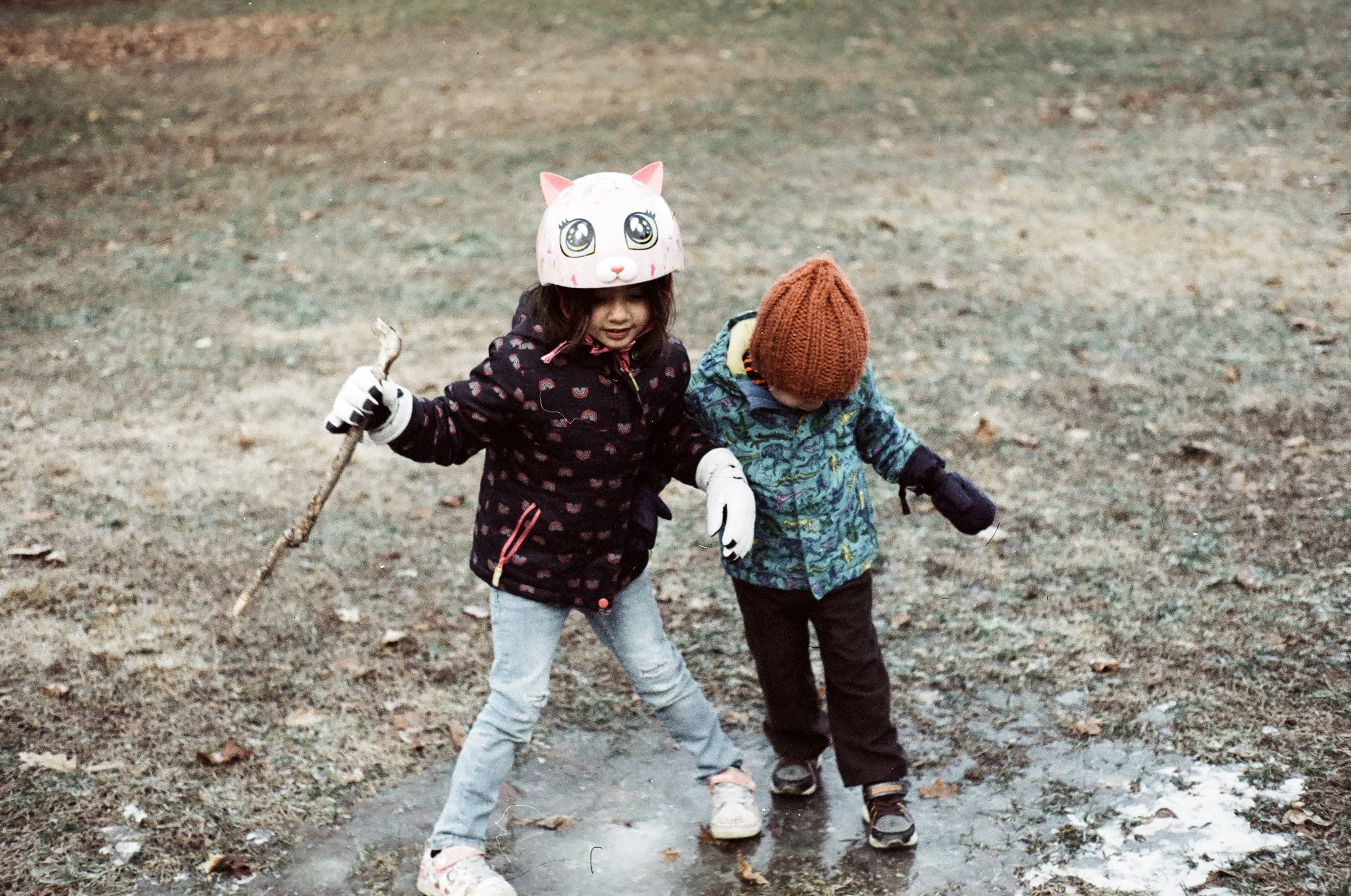 two small children in winter coats slip around on a patch of ice in a field. the child on the left is wearing a pink cat helmet and black coat with rainbows on it. the child on the right is wearing an orange knit hat, a blue coat and snow pants.