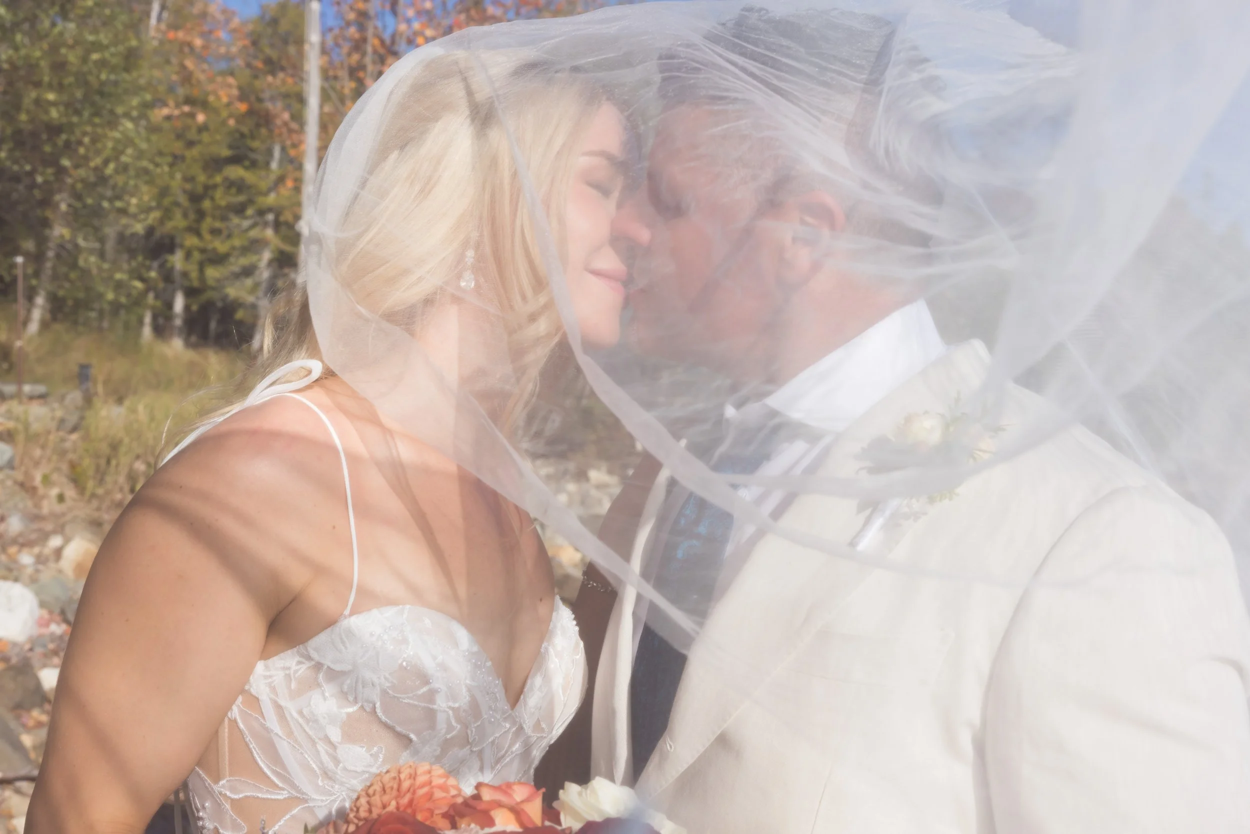 an images of a blonde bride and a groom in a cream tuxedo kissing under a veil. Their eyes are closed.  The veil is being swept to the right by the wind