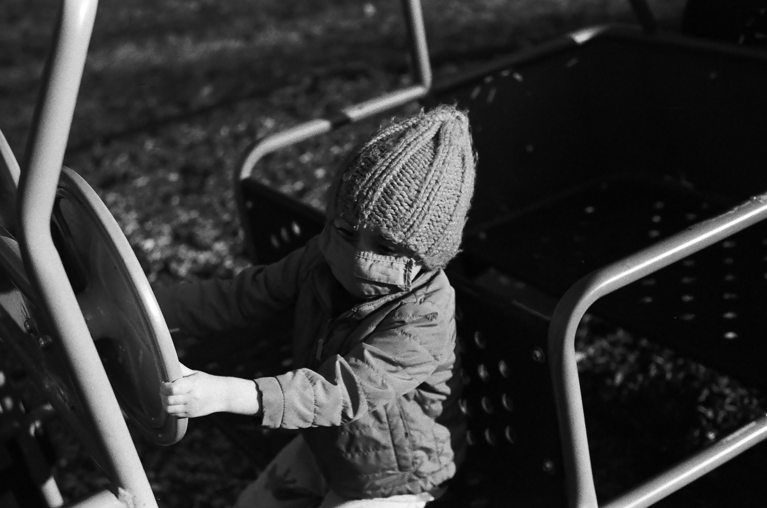 an image of a child with a mask on, a jacket, and a hat, driving a play car on a playground
