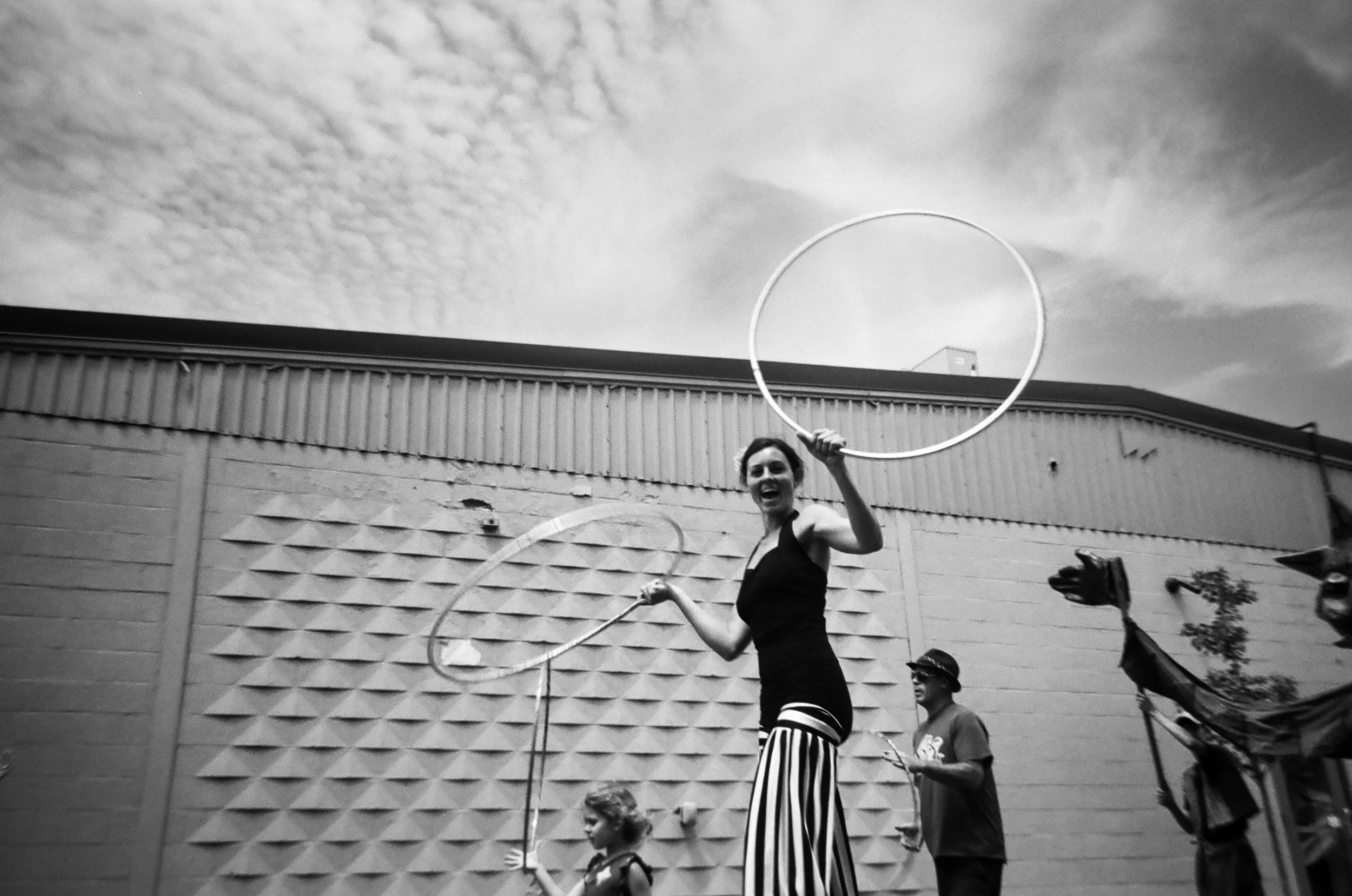 a woman on stilts wearing a black leotard, striped pants and carrying two hula hoops walks by.