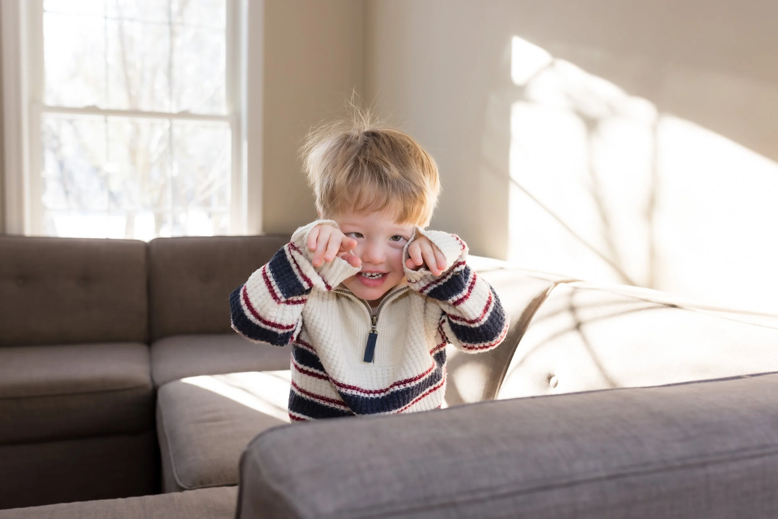 A blond haired child in a striped sweater reveals his face. There are shadows on the walls behind him from light coming through a window. His hair is messy and standing on end.