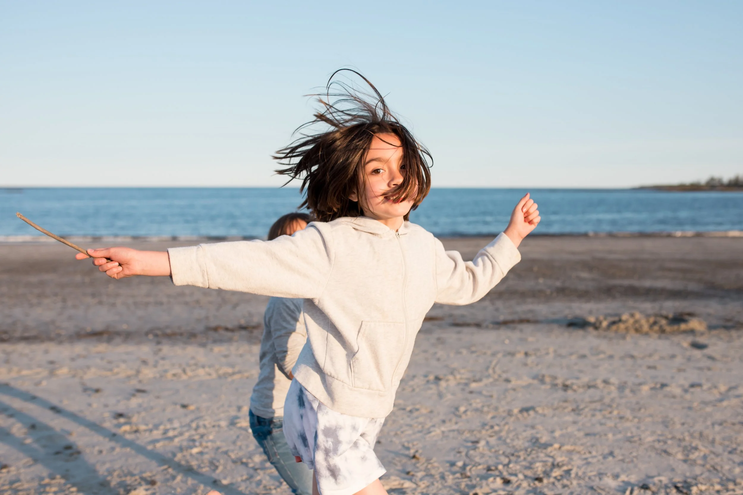 an image of a child running on the beach, looking at the camera, with a stick in one hand outstretched behind them. The sky is blue. they are wearing a light gray hooded sweatshirt. Their hair is tousled in the wind.