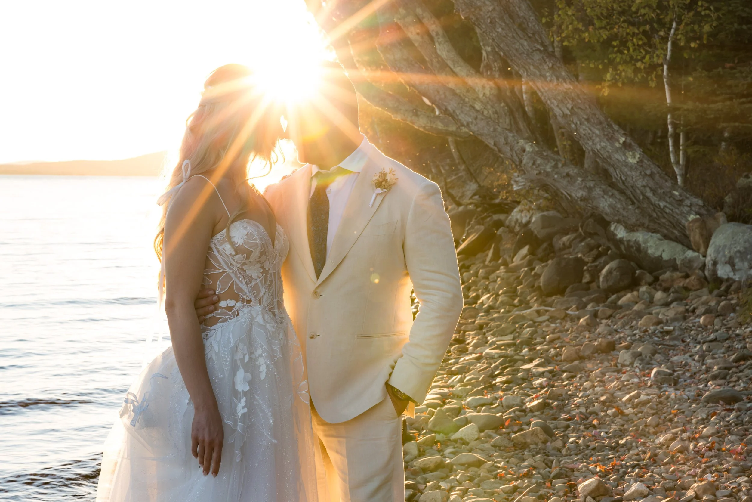 an image of a bride and groom, foreheads pressed together. they are standing on a rocky lake shore. There is a starburst of sunbeams streaming through right where their heads connect.