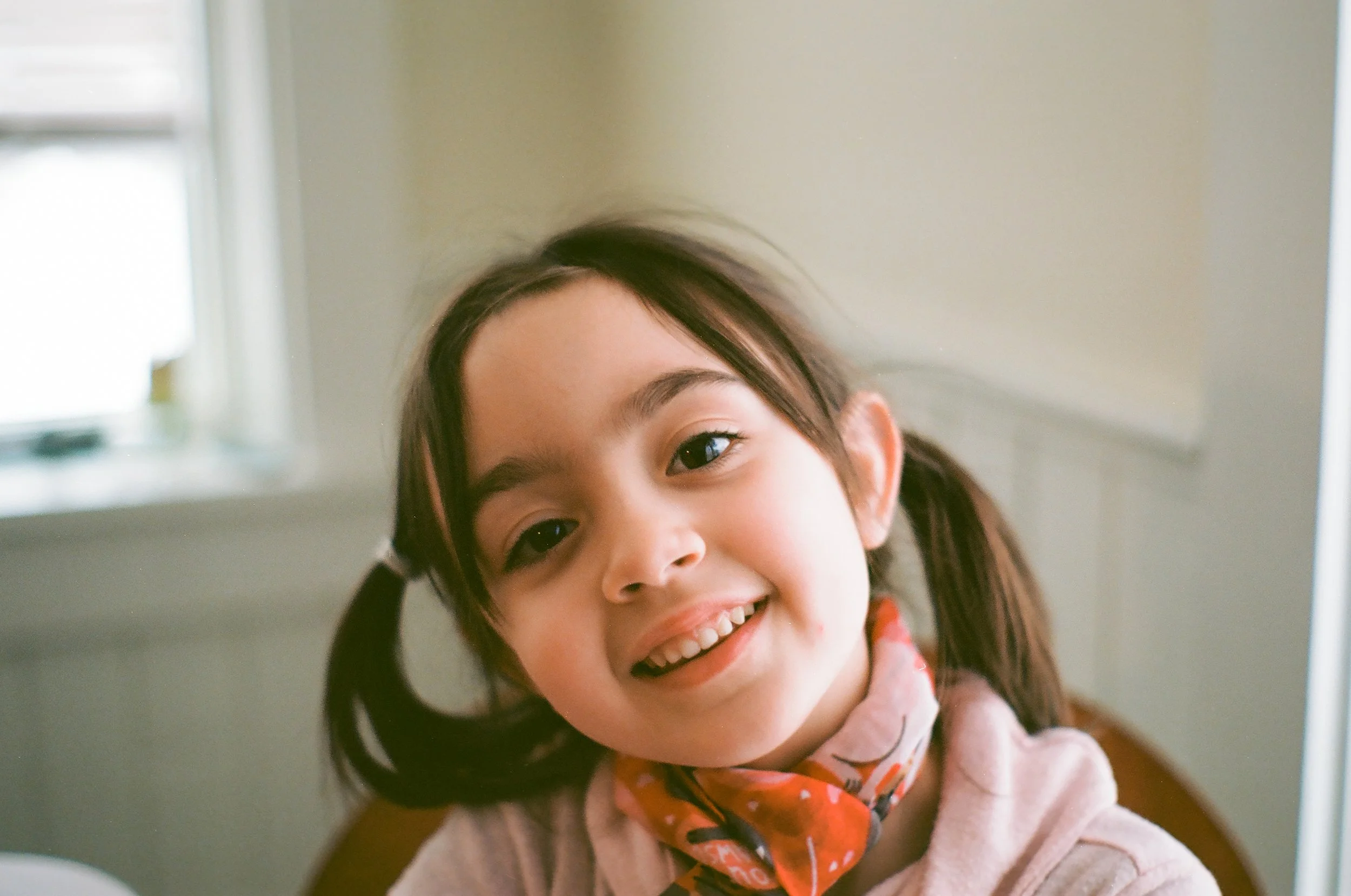 a girl with brown hair in pigtails, wearing a pink sweater sits in a chair and smiles for the camera