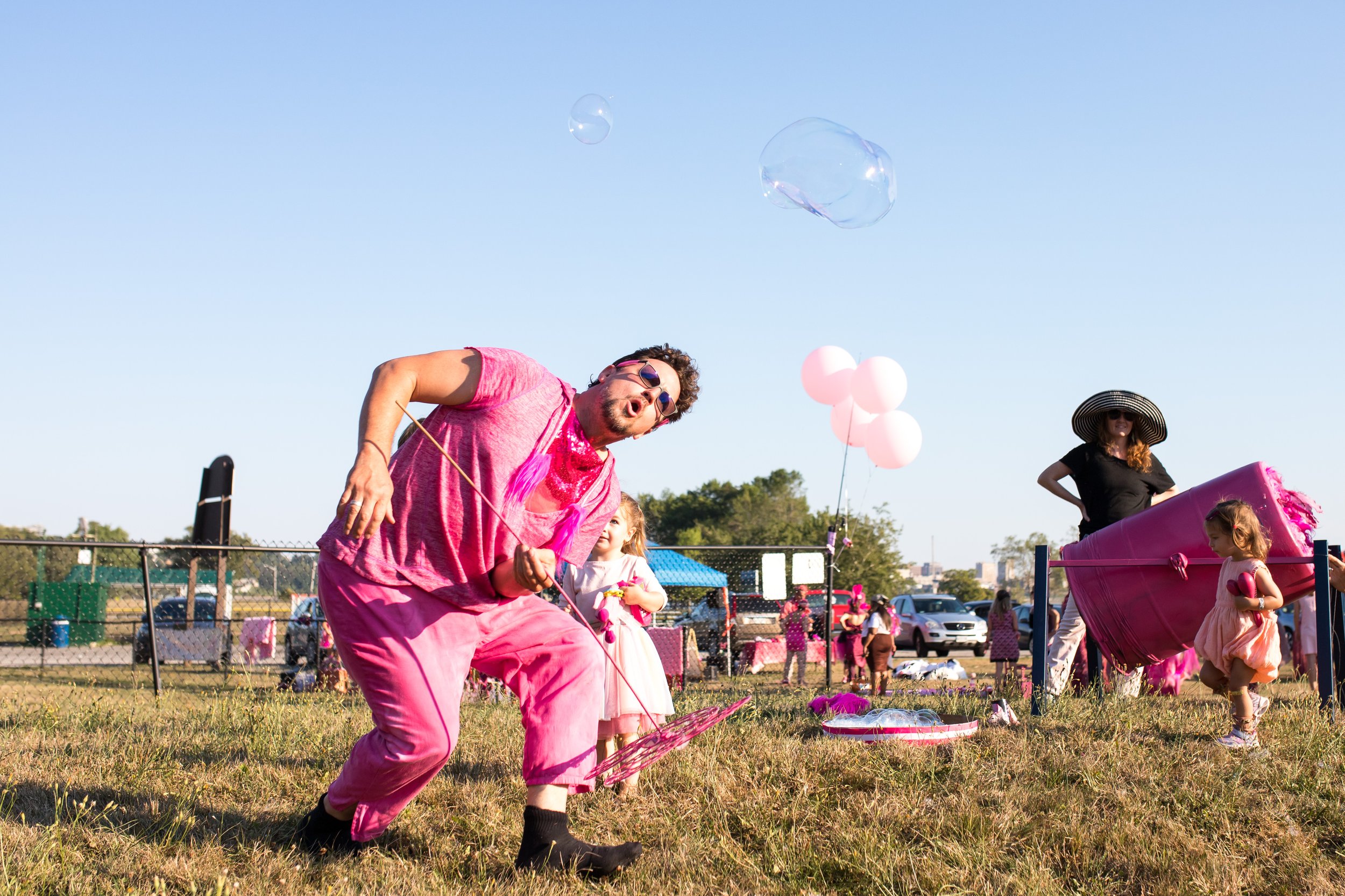 an image of a man dressed in pink blowing giant bubbles. the sky is blue, there is a large crowd of people all dressed in pink. there is. a woman in a black and white outfit looking , there are pink balloons