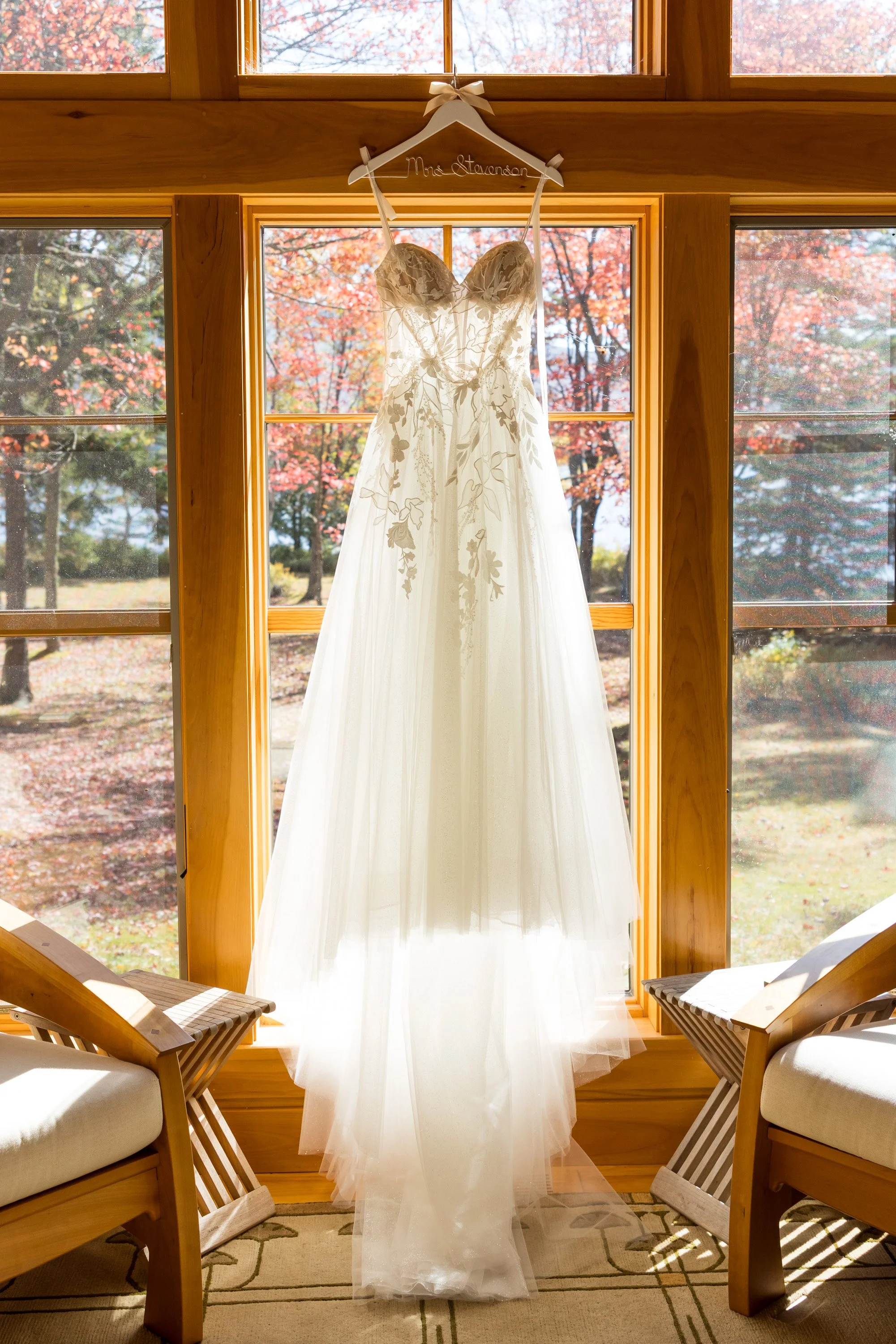 a wedding dress hangs in a tall window, the trees outside have fall colors. The dress is white, with Lacey flowers and ribbons.