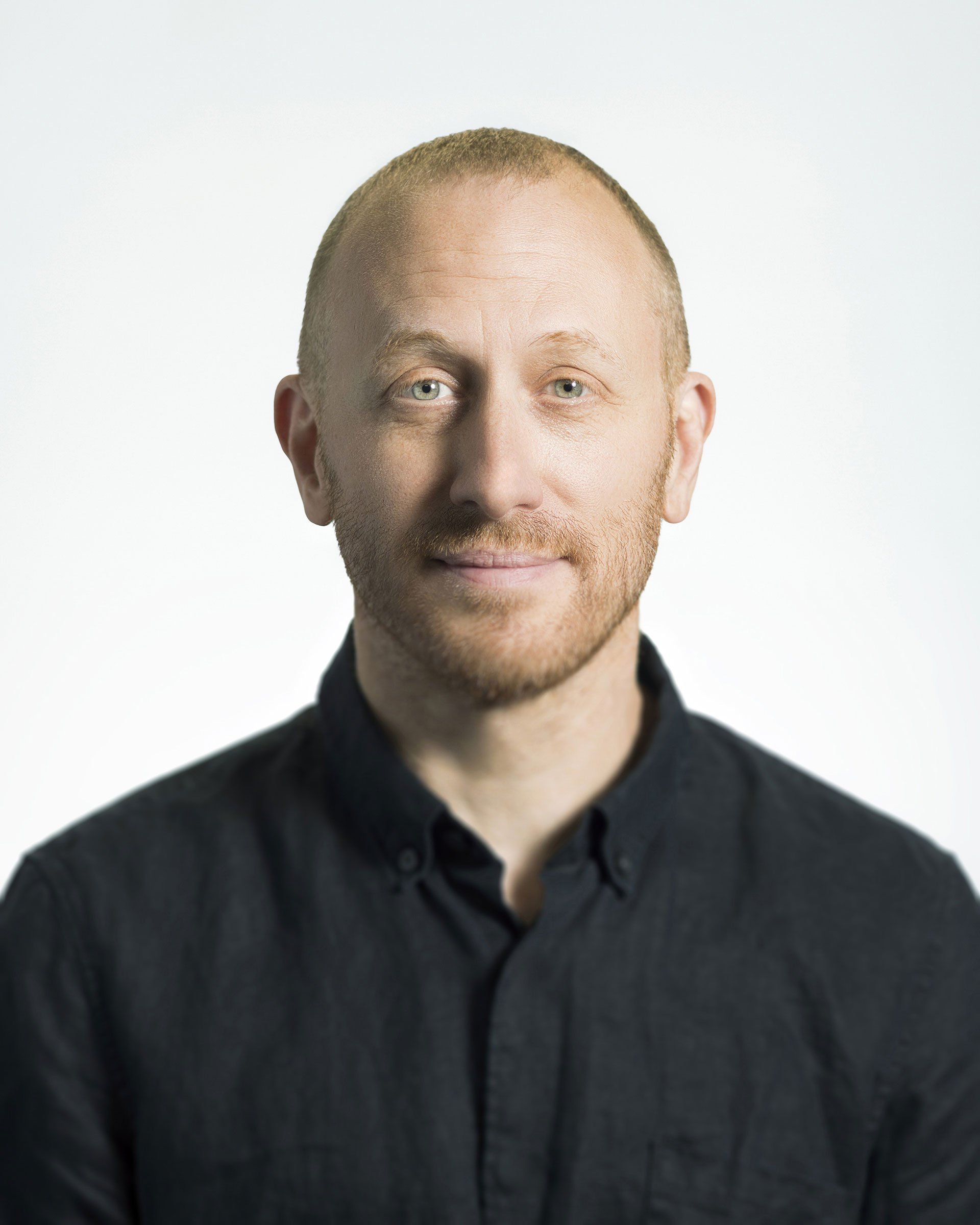 Headshot of a man with short, light-colored hair and a beard, wearing a black collared shirt, smiling slightly against a white background.