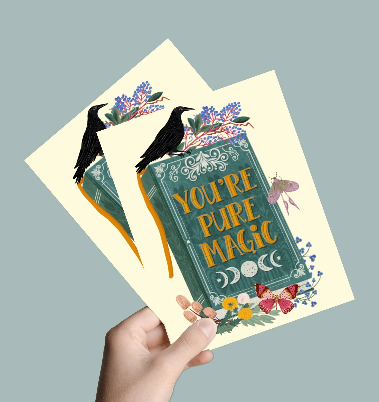 hand holding two crow and magic book friendship cards on a soft green background
