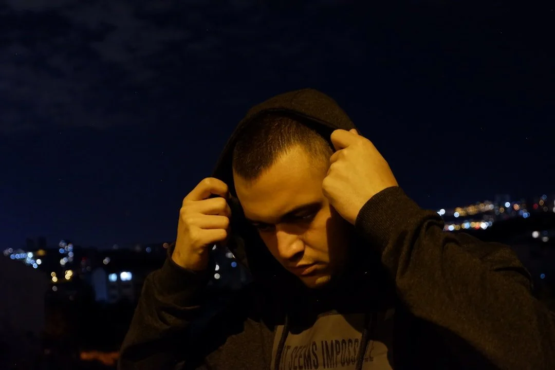 A young man with short hair wearing a dark hoodie, adjusting the hood at night with city lights in the background.