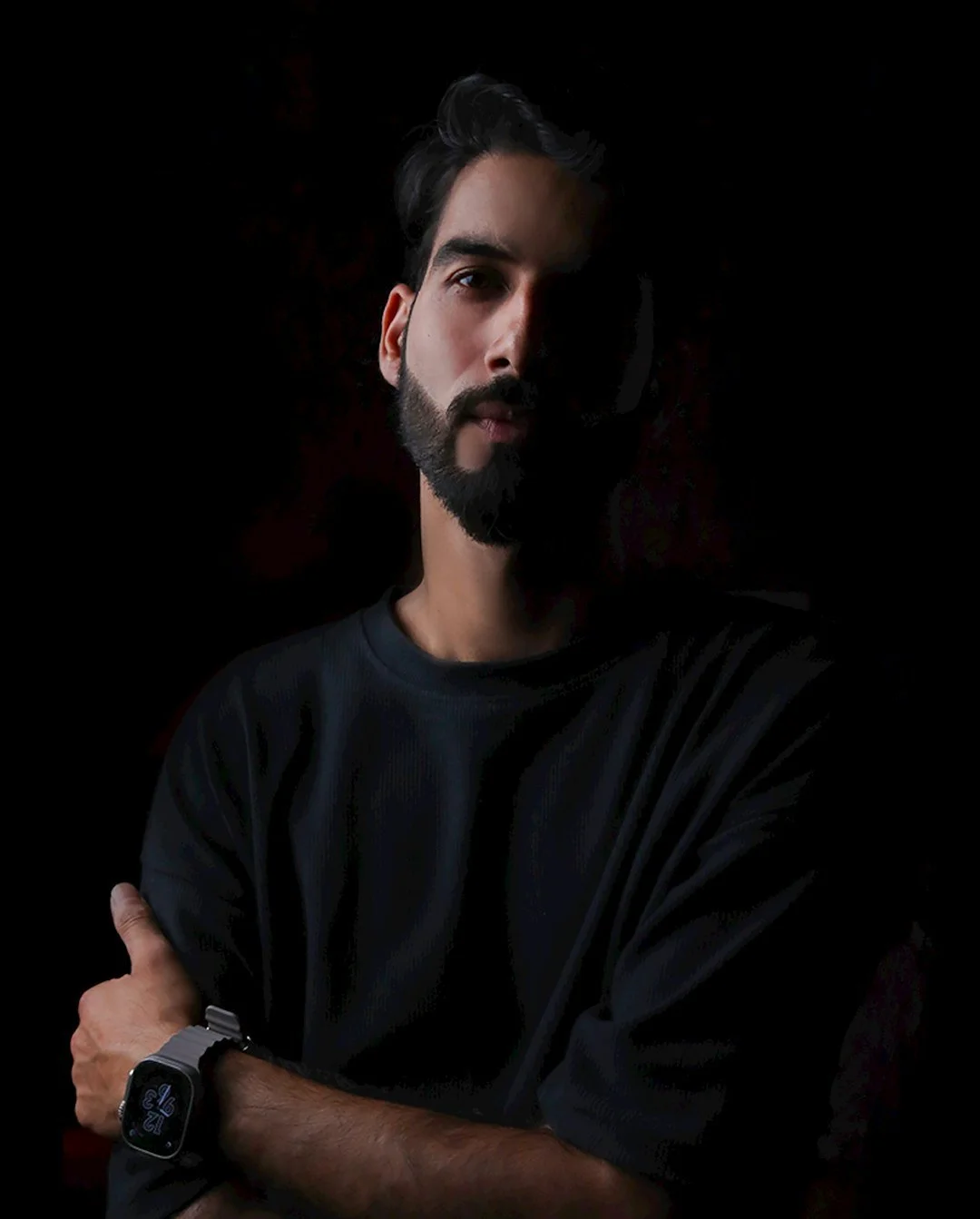 A young man with dark hair and a beard, wearing a black shirt and a smartwatch, standing against a dark background.