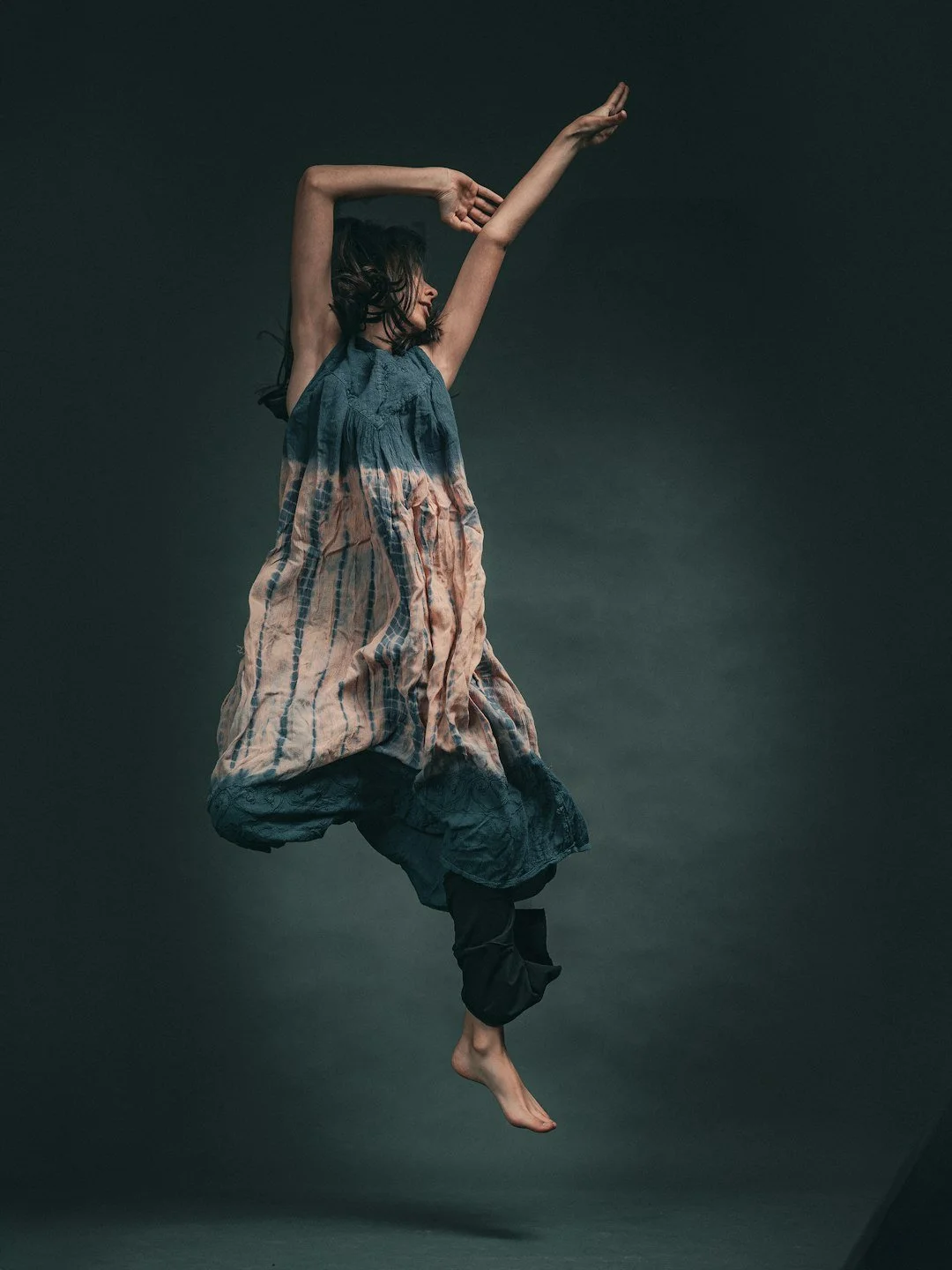 A woman dancing, with one arm raised high and the other bent, wearing a loose, multicolored dress and black pants, set against a dark background.