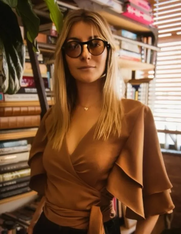 A woman with long blonde hair wearing glasses and a tan wrap-around top stands in front of a bookshelf filled with books and plants, with sunlight coming through window blinds.