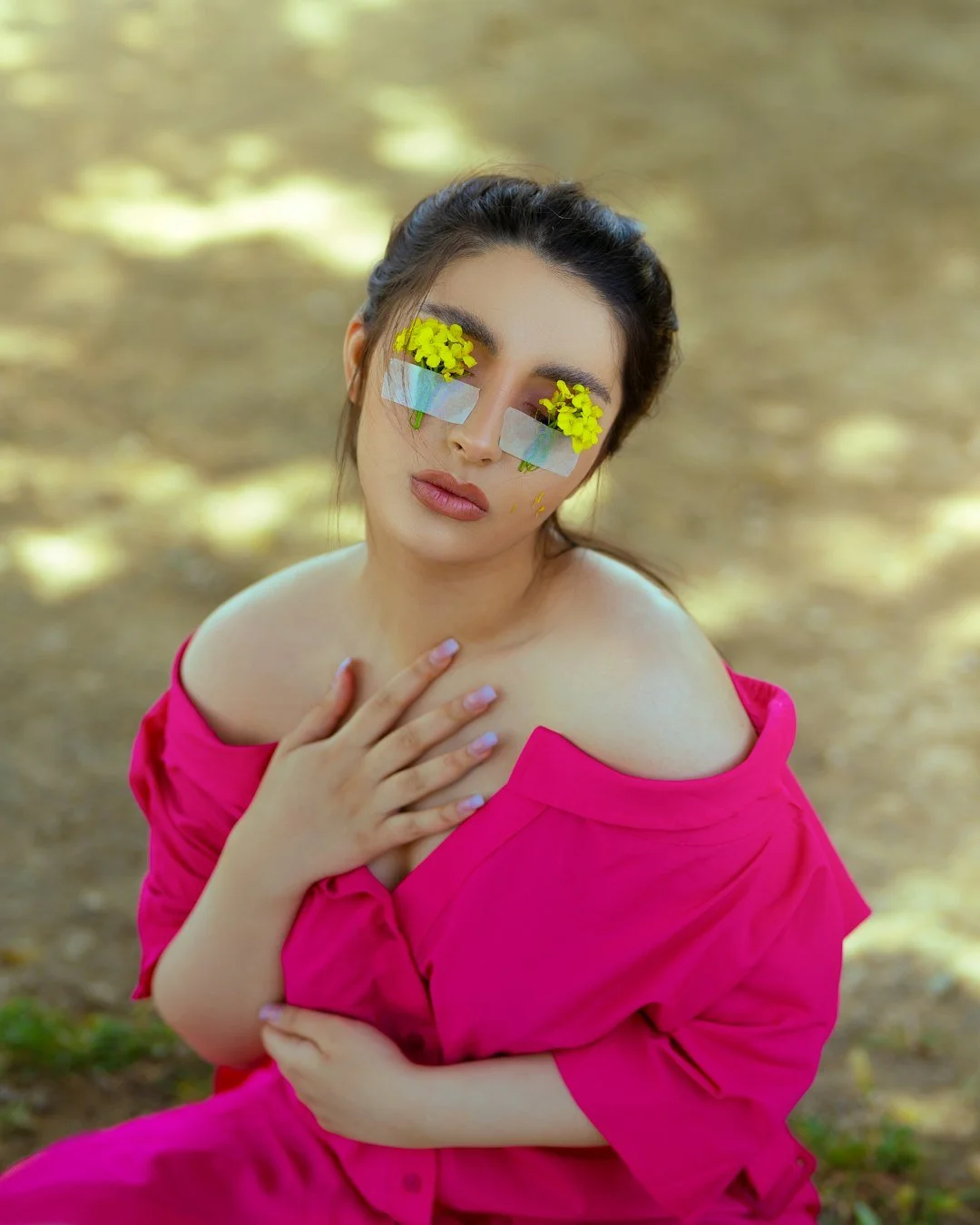 A woman with flowers on her eyelids and tape on her cheeks in a pink off-shoulder dress, posing outdoors with her hand on her chest.