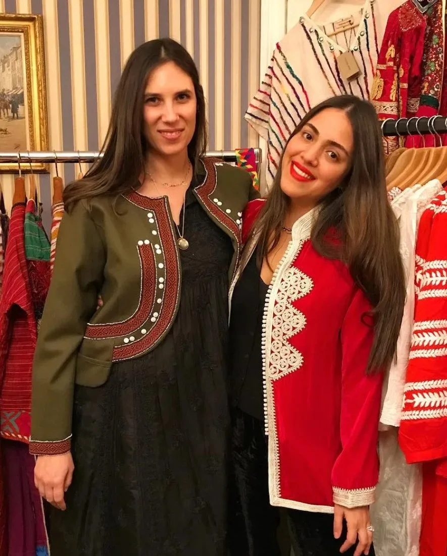 Two women standing inside a clothing store, surrounded by racks of traditional and festive clothing, smiling at the camera.