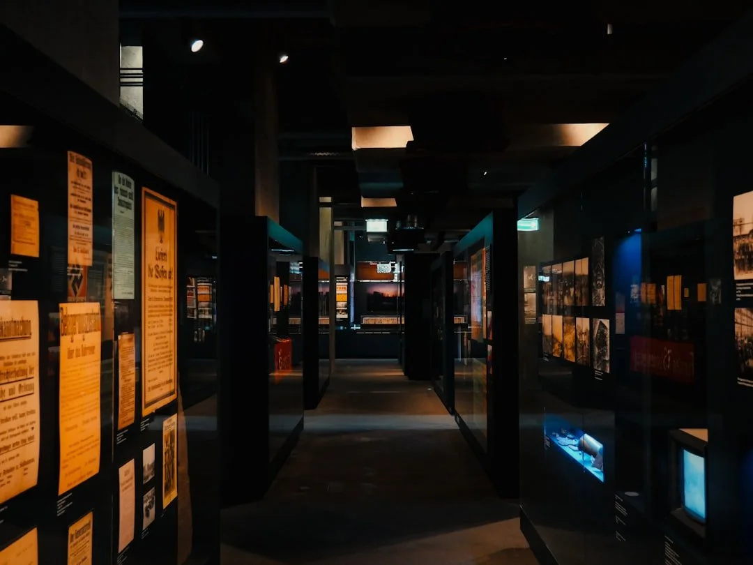 Dimly lit museum exhibit with black panels displaying photographs and informational panels, with a sunset visible through windows in the background.