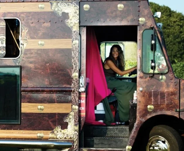 A woman sitting inside a creatively decorated food truck with a red curtain, smiling and holding the steering wheel.