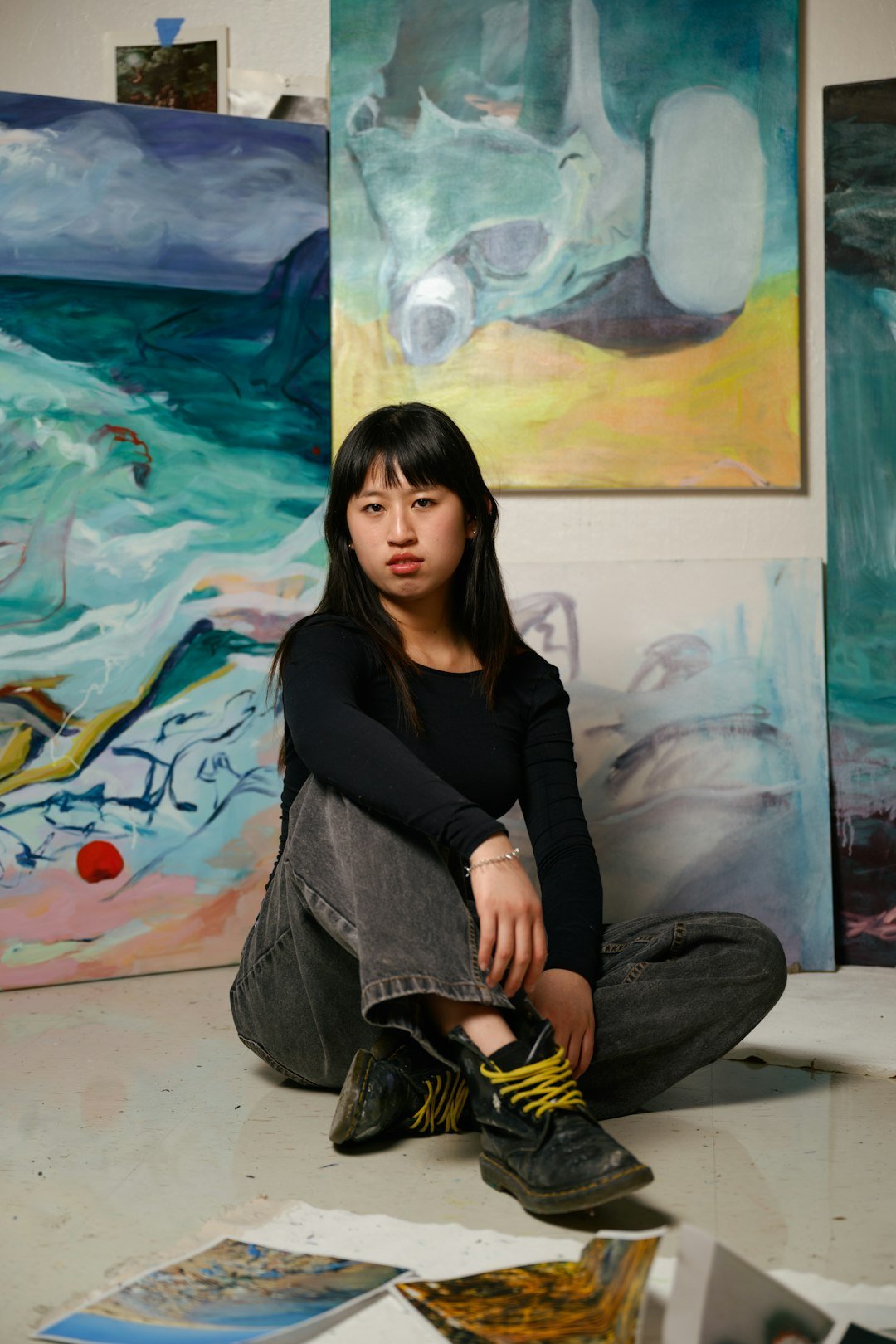 A young woman with long black hair sitting on the floor in front of large, colorful abstract paintings. She is wearing a black long-sleeve shirt, gray jeans, and black boots with yellow laces. There are scattered photographs or prints on the floor around her.