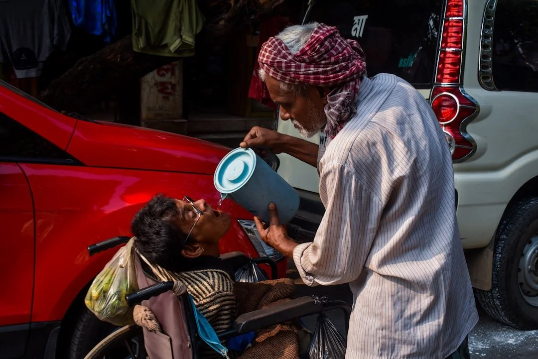 An elderly man with a headscarf offers water to a woman in a wheelchair on a street with parked cars.