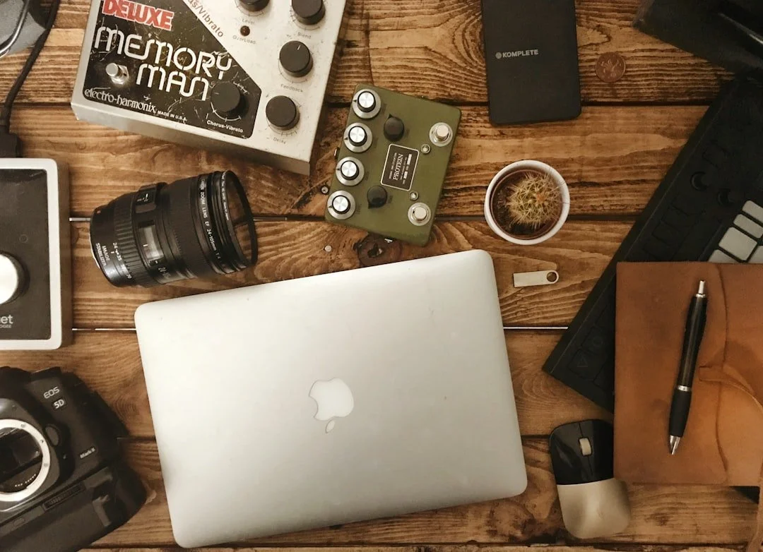 A flat lay of electronic devices and accessories on a wooden surface, including a closed MacBook, a camera, a camera lens, a MIDI controller, a keyboard, a mouse, a pen, a small potted cactus, a cup of coffee, and a music effect pedal.