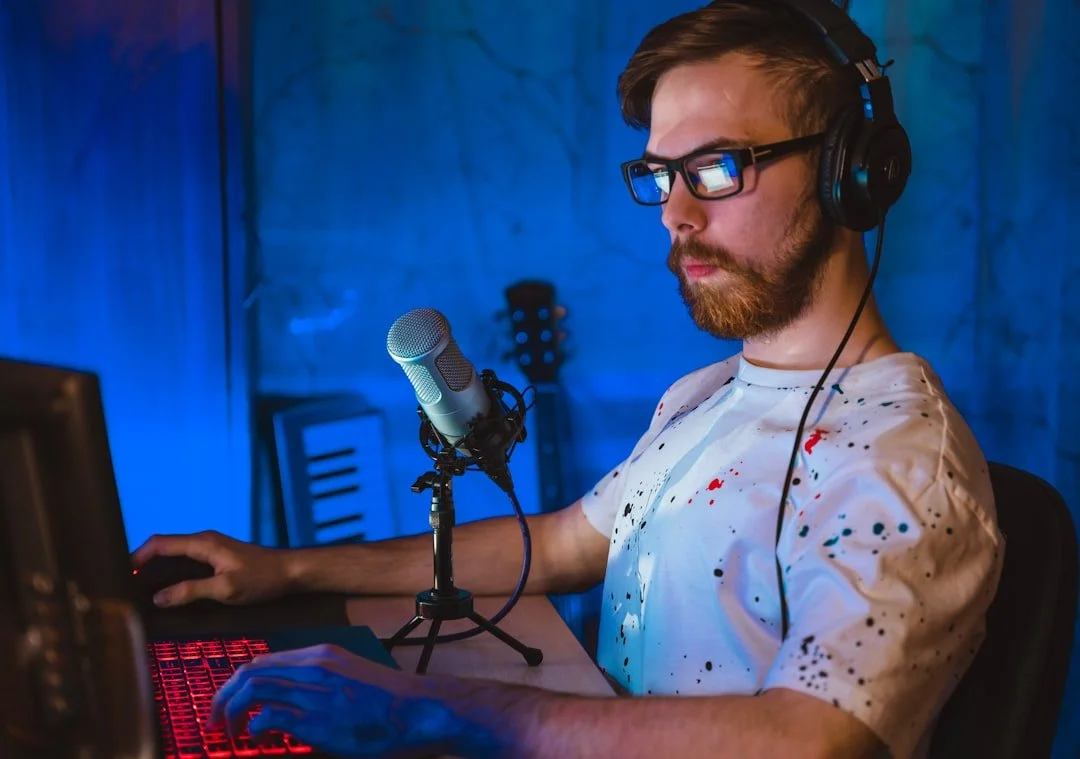 Man with glasses and headphones working at a computer with a microphone in a dimly lit room.