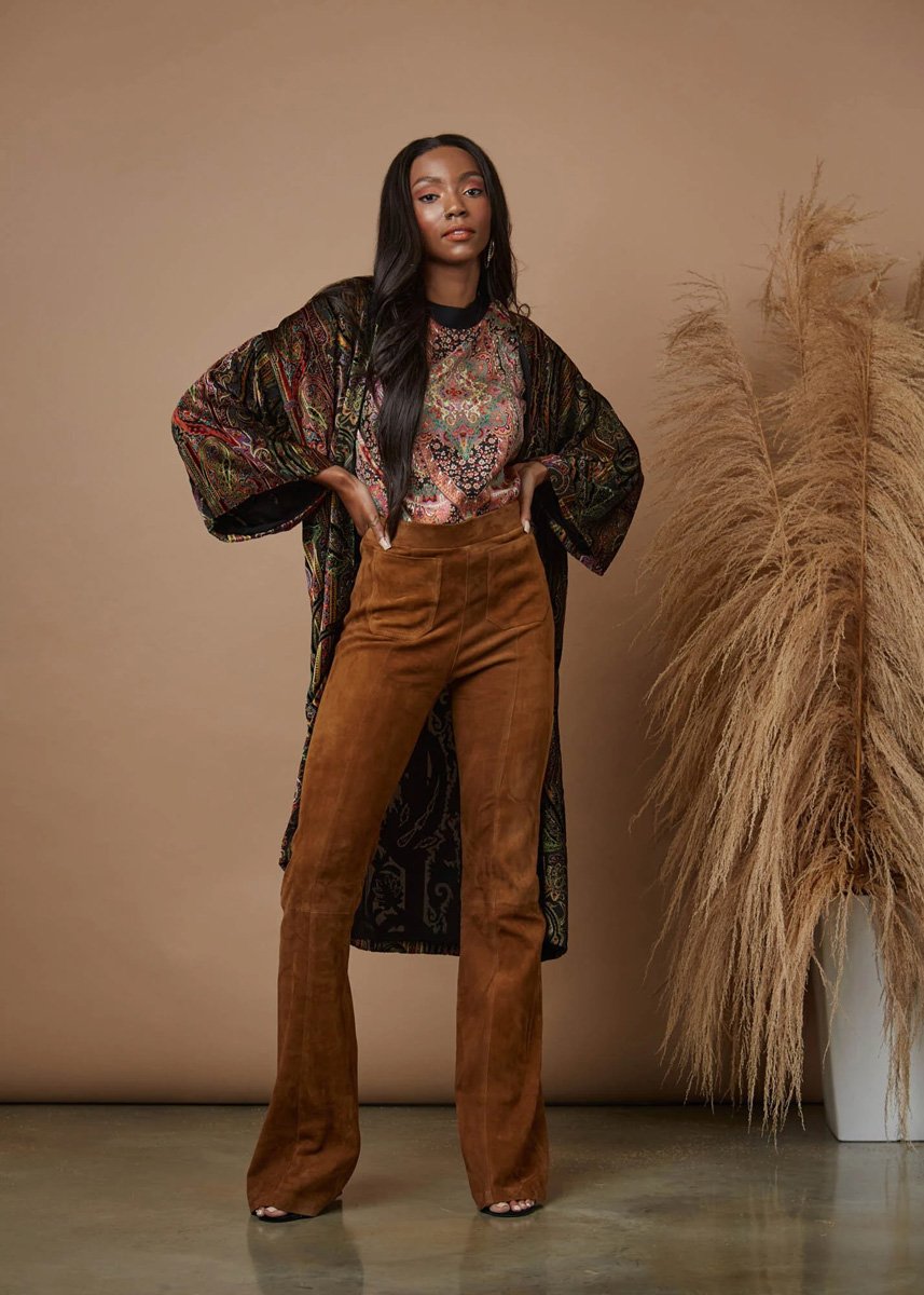A woman standing with her hands on her hips, wearing a patterned blouse, a long dark kimono, and brown flared pants, posed against a neutral beige background with tall ornamental grasses.