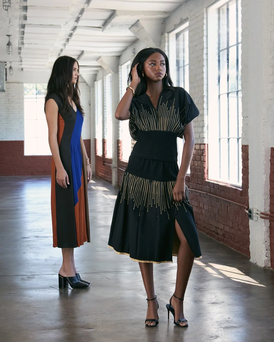 Two women in stylish dresses standing in a spacious, well-lit industrial-style room with large windows and brick walls. One woman in the foreground is wearing a black dress with gold accents and high heels, while the other woman in the background is 