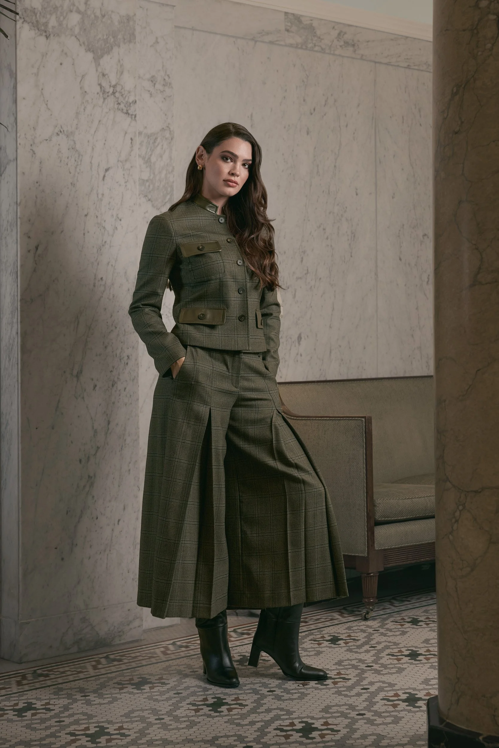 Two women posing indoors with a stone wall and a beige fabric backdrop behind them. One woman with long dark hair, wearing a beige sweater, black leather pants, and black heeled boots, sitting on a wooden chair. The other woman with long wavy brown hair, wearing a dark denim shirt, a tan cardigan draped over her shoulders, dark jeans, and tan heeled boots, standing beside her.
