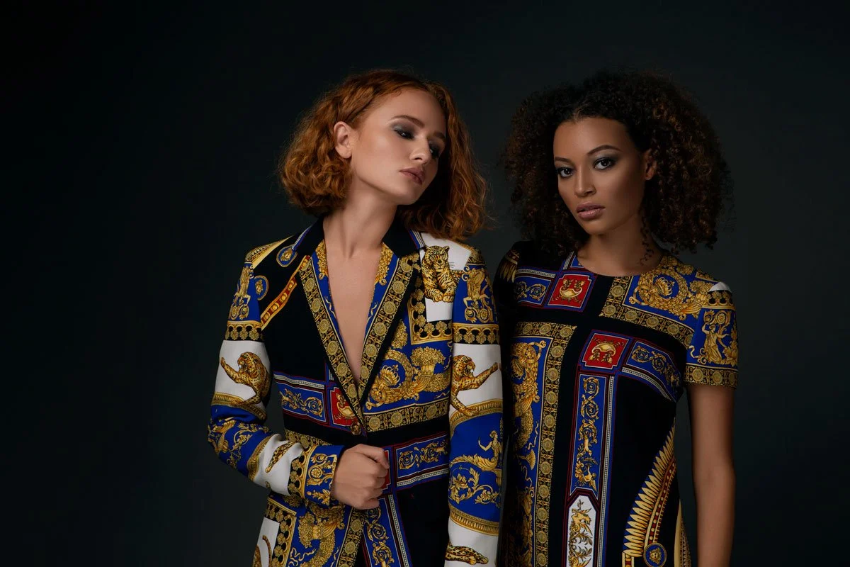 Two women with curly hair wearing ornate, patterned jackets standing against a dark background.
