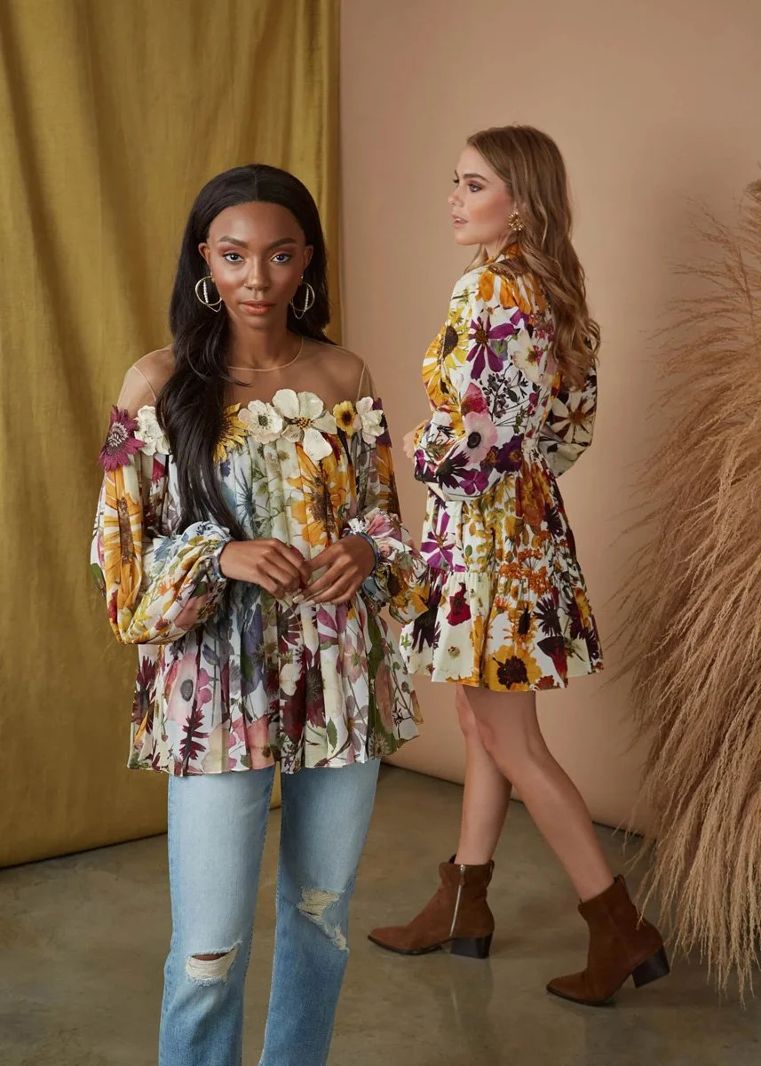 Two women wearing floral patterned outfits with earthy tones, one standing in front wearing a long-sleeve top with a floral design and ripped jeans, the other in the background wearing a floral dress with ruffles, both standing indoors with mustard-c