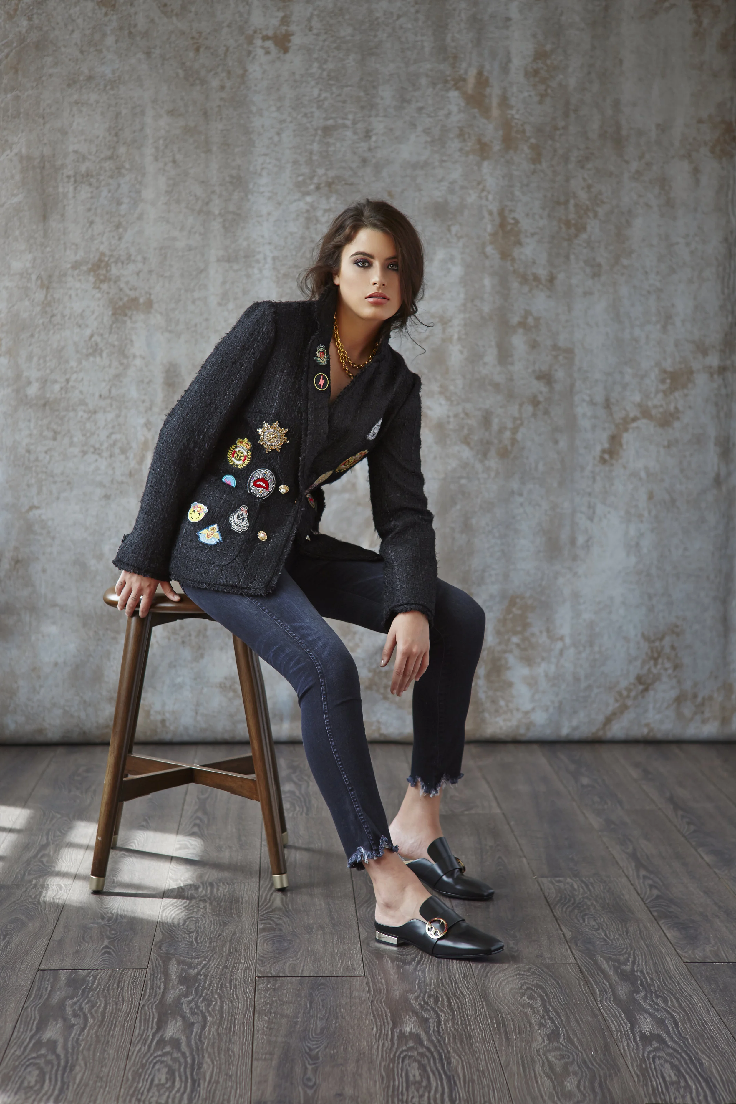 A woman sitting on a wooden stool against a textured gray wall. She has dark wavy hair, wears a black blazer with colorful patches, dark jeans with frayed hems, and black slip-on shoes.