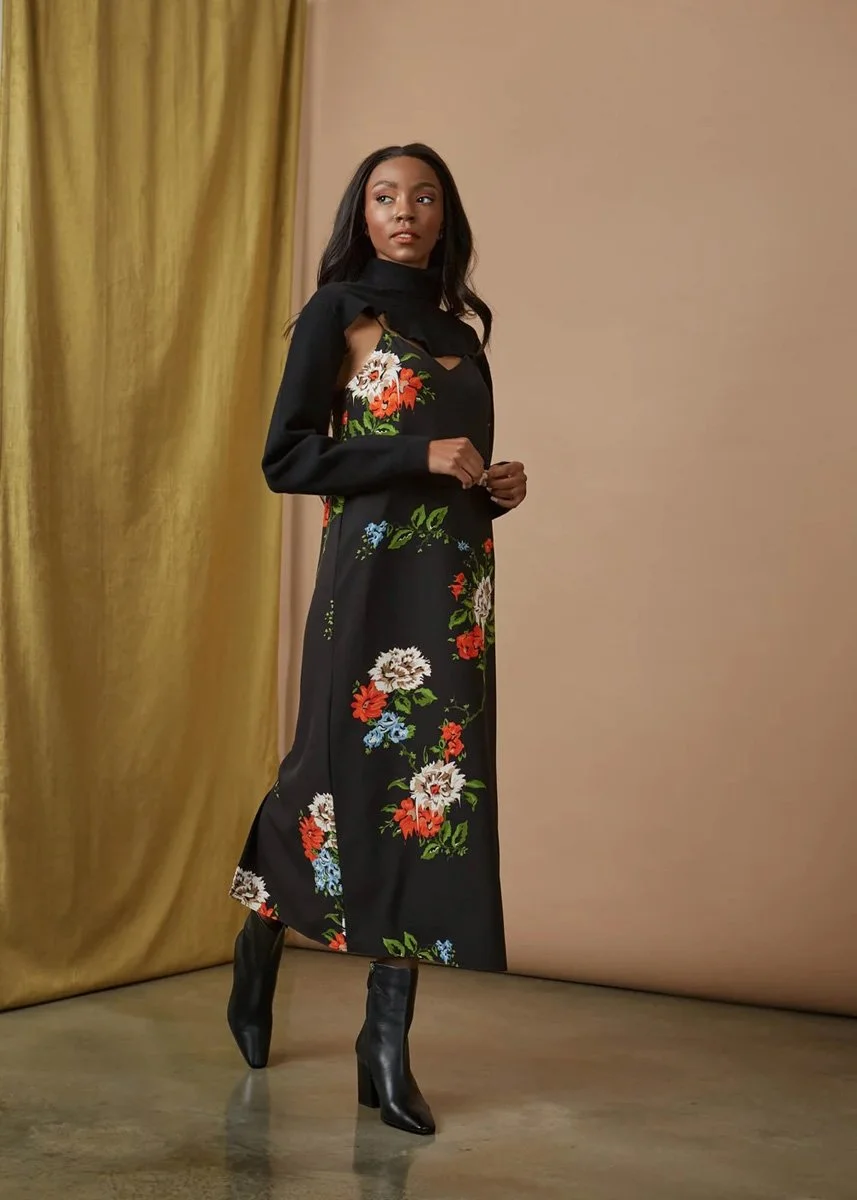 A woman wearing a black dress with colorful floral embroidery, paired with black heeled boots, standing indoors against beige and gold curtains.