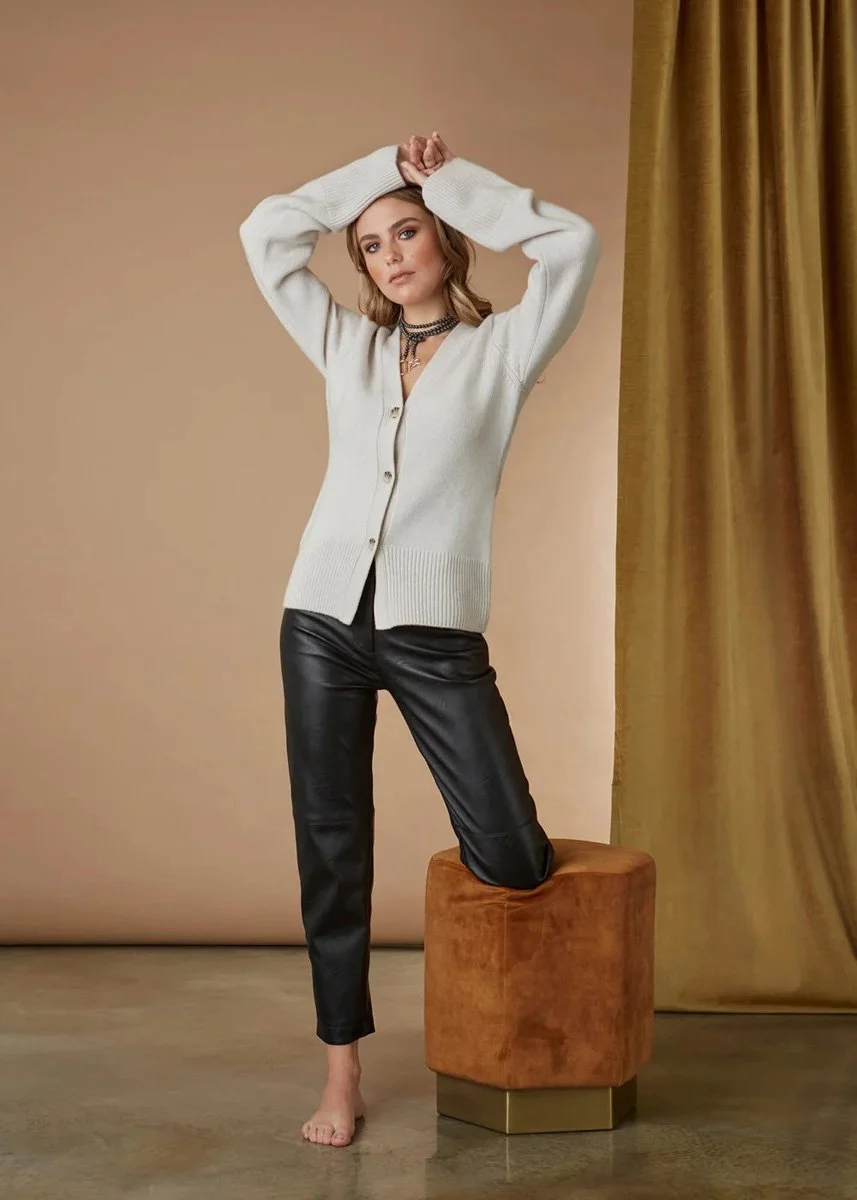 A woman with shoulder-length brown hair posing with one knee on a small orange stool. She is wearing a cream-colored cardigan, black leather pants, and layered necklaces, against a beige background with a gold curtain on the right side.