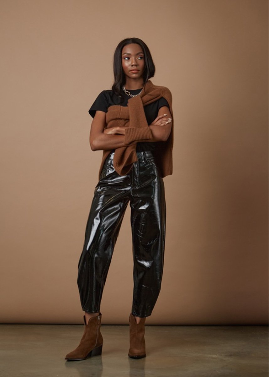Young woman standing against a beige background, wearing black shiny leather pants, a black t-shirt, brown ankle boots, and a brown sweater draped over her shoulders, with arms crossed and a neutral expression.