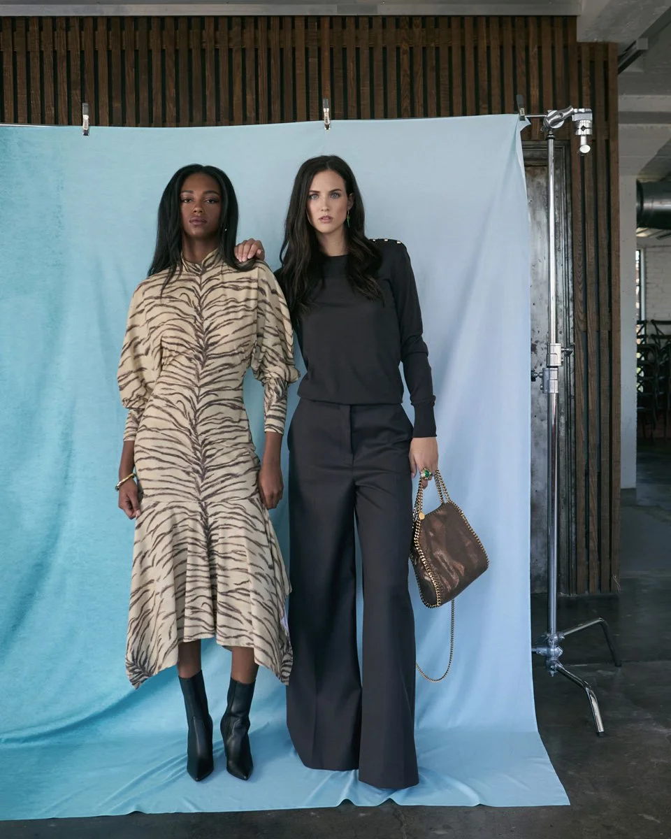 Two women posing in fashionable outfits in front of a light blue backdrop, with one wearing a beige and black zebra print dress and the other in a black top and wide-leg pants, holding a brown handbag.