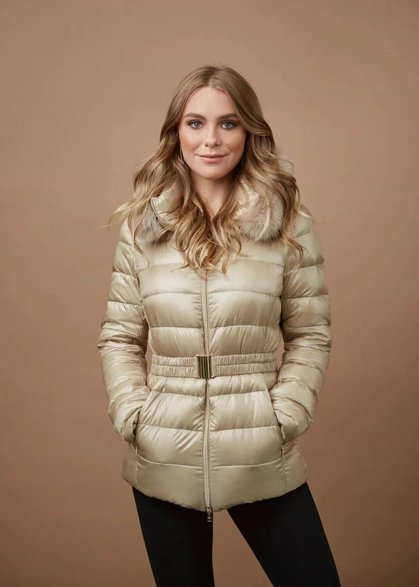 A woman with wavy blonde hair wearing a beige puffer jacket with a fur collar, standing against a plain brown background.